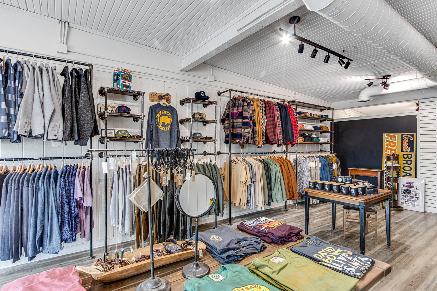 Image of the inside of the Everywear Menswear and Lifestyle store located in Rockford Michigan. The photo shows a table in the foreground with t-shirts and bracelets and the background shows racks of clothing.