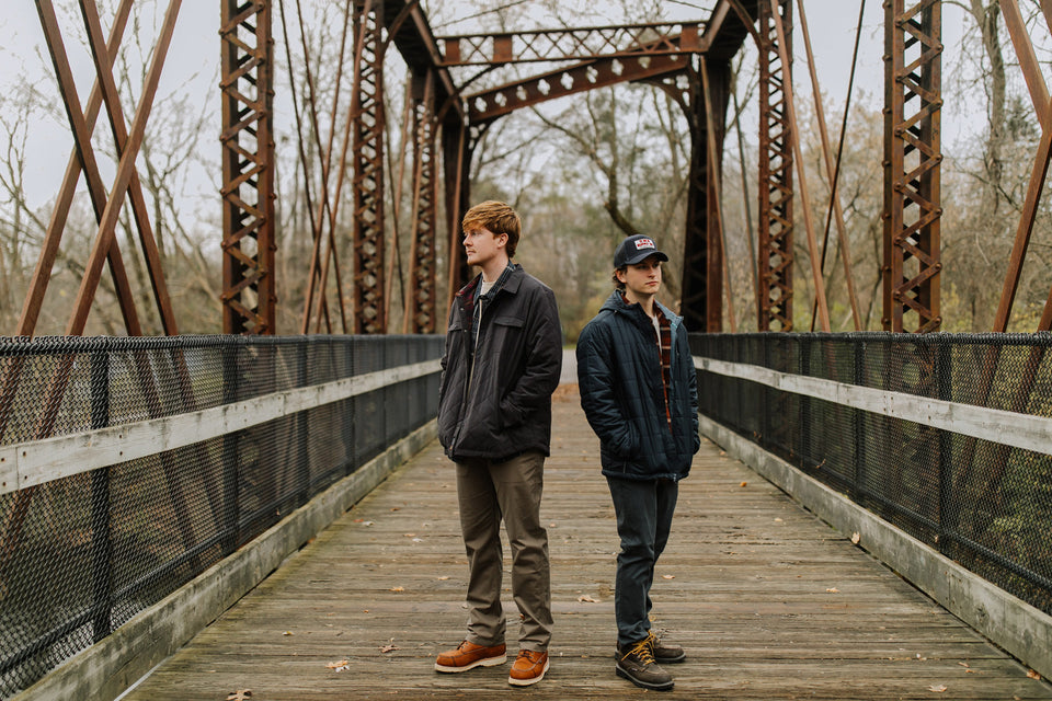 Two guys standing in the middle of a bridge in fall weather. They're wearing rugged and outdoor style clothing from Everywear.